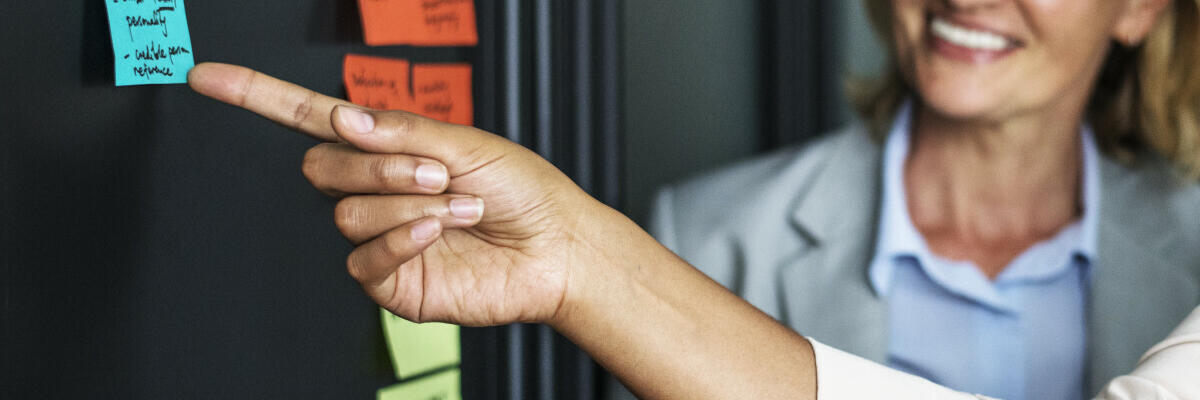 Eine Frau schaut auf eine Memo-Wand mit Notizzetteln darauf und eine andere Person zeit auf einen Notizzettel.