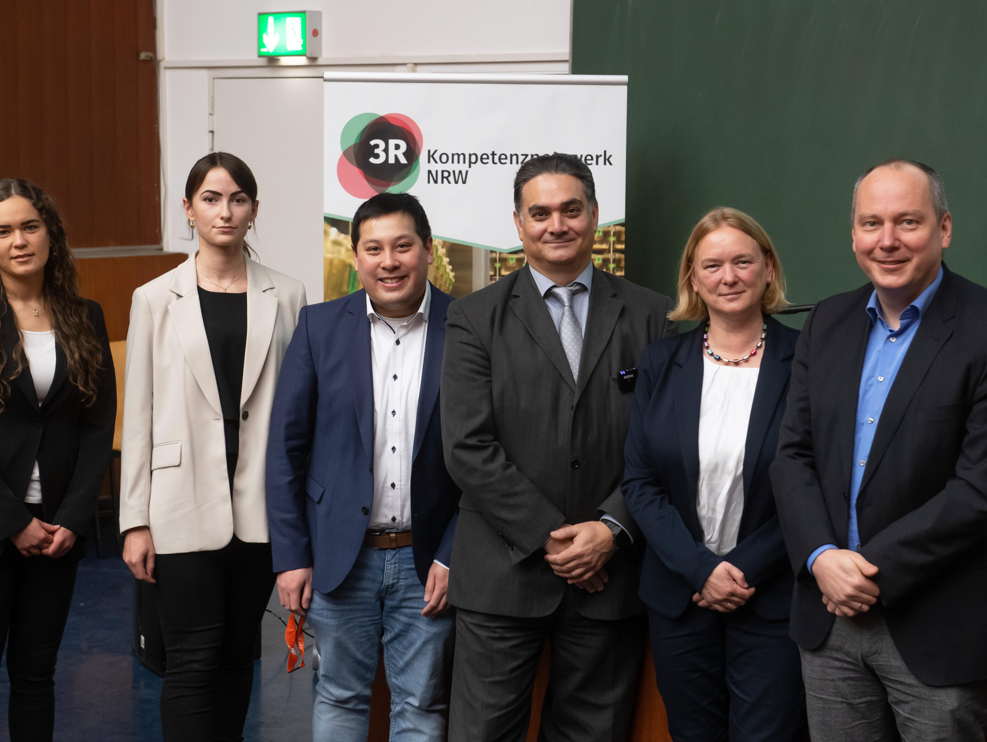 Gruppe von sechs Menschen stehen nebeneinander in einem Hörsaal vor einem Pult und lächeln in die Kamera