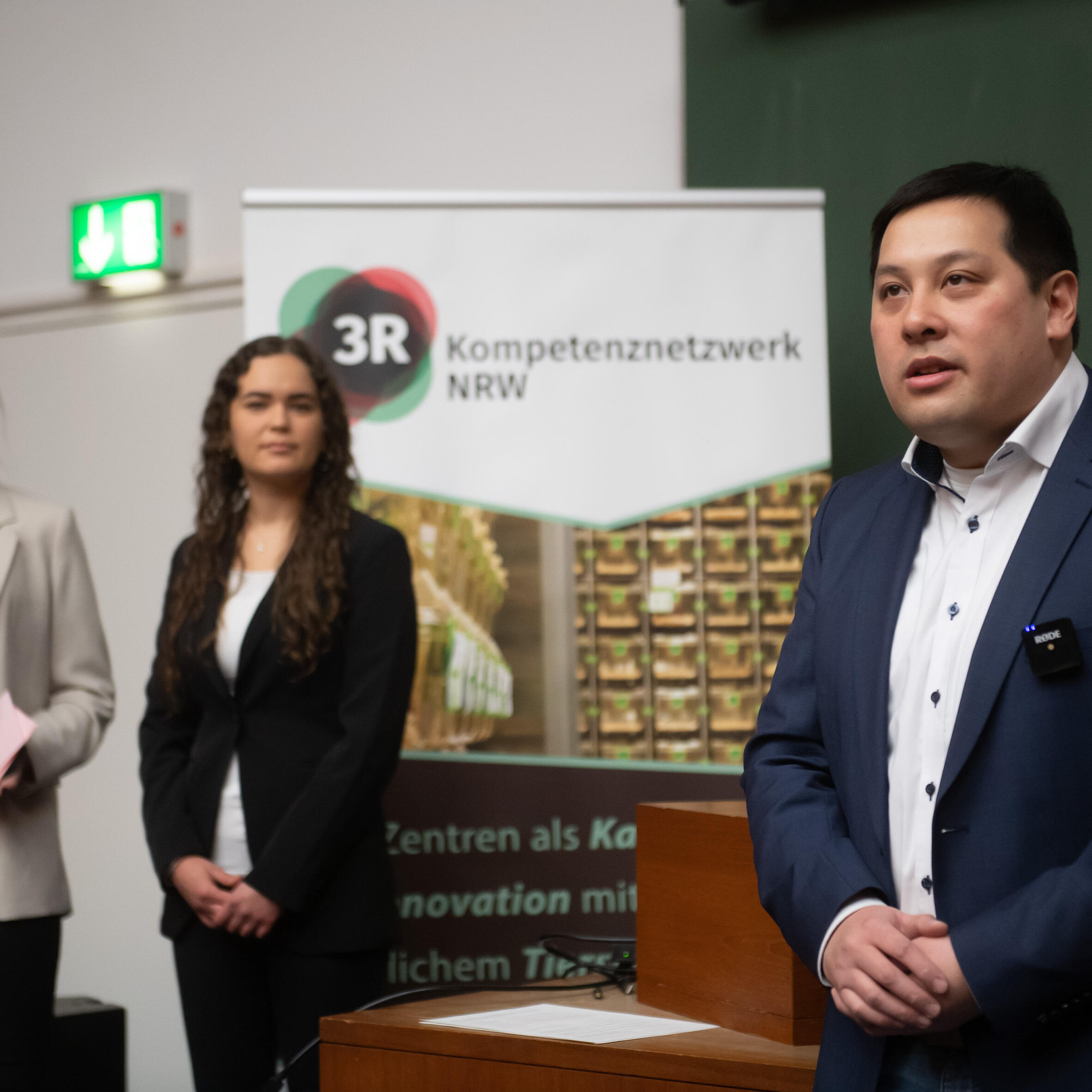 Die 3R-Geschäftsstelle stellt sich vor: Mann mit weißen Hemd und blauen Sakko steht im Vordergrund eines Hörssaals und spricht mit gefalteten Händen. Im Hintergrund stehen zwei Frauen in einem beigen Balzer mit schwarzen Oberteil und einem schwarzen Blazer mit hellen Oberteil.
