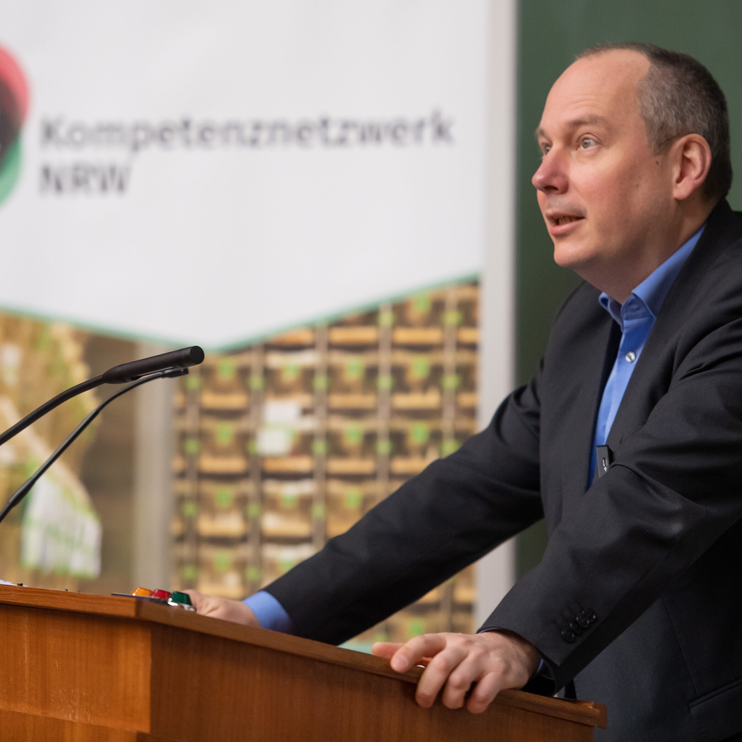 Grußwort Dekan Prof. Weber: Mann im schwarzgrauen Anzug mit hellblauem Hemd hält mit Blick ins Publikum eine Rede am Rednerpult in einem Hörsaal