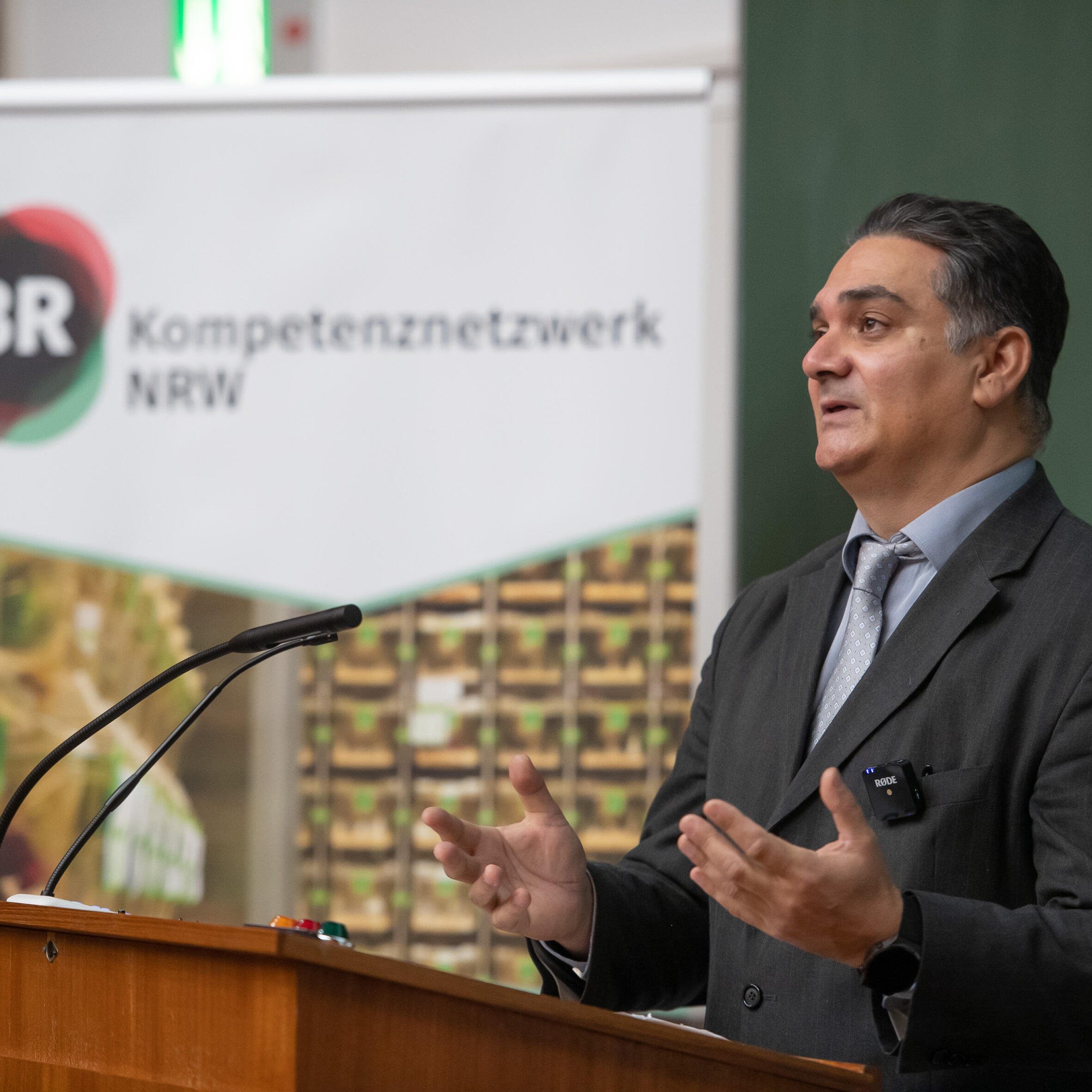 Grußwort Prof. Tolba: Mann in einem schwarzgrauen Anzug mit hellgrauem Hemd und Krawatte hält am Rednerpult eines Hörsaals eine Rede mit Blick ins Publikum und beide Hände sind angehoben