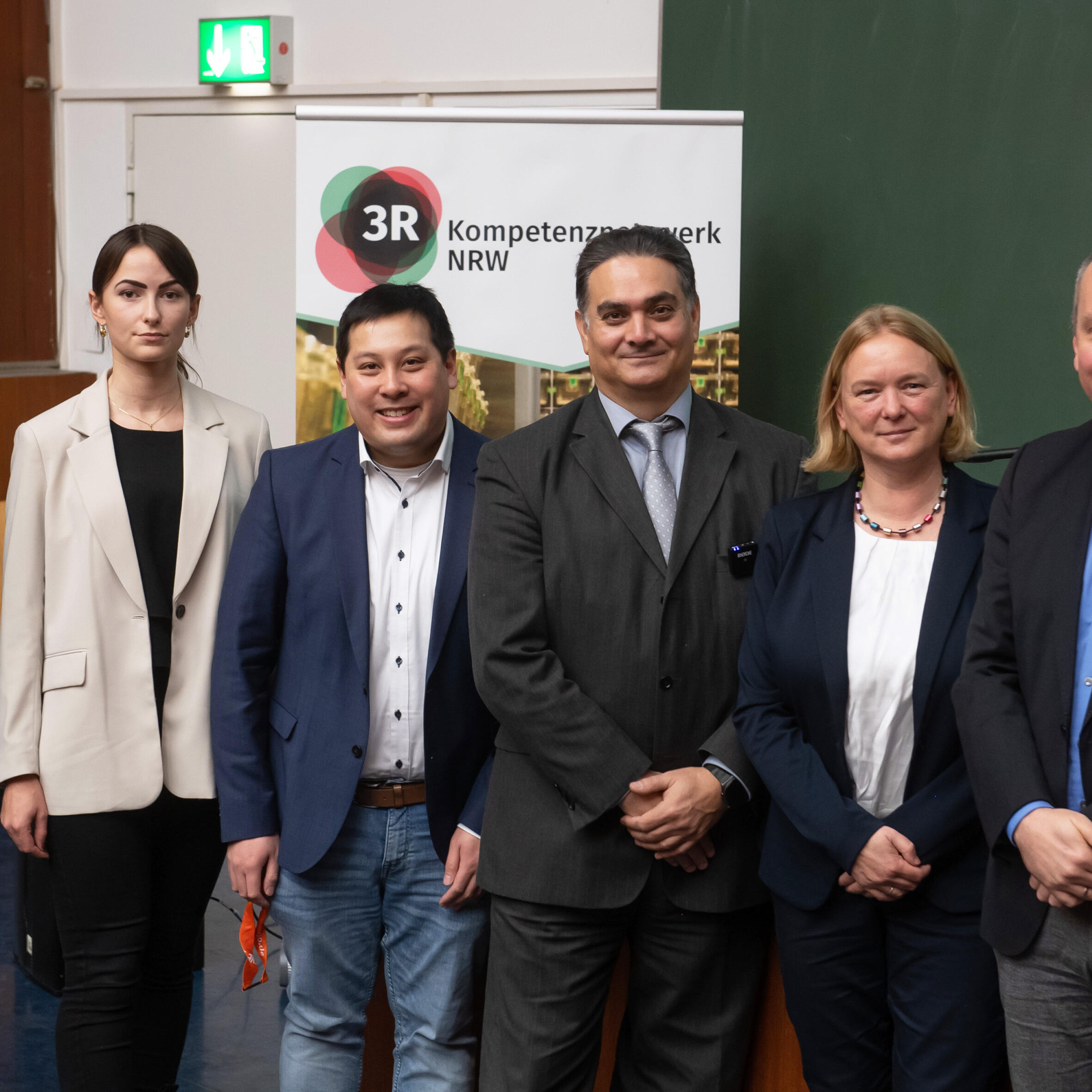 Gruppe von sechs Menschen stehen nebeneinander in einem Hörsaal vor einem Pult und lächeln in die Kamera