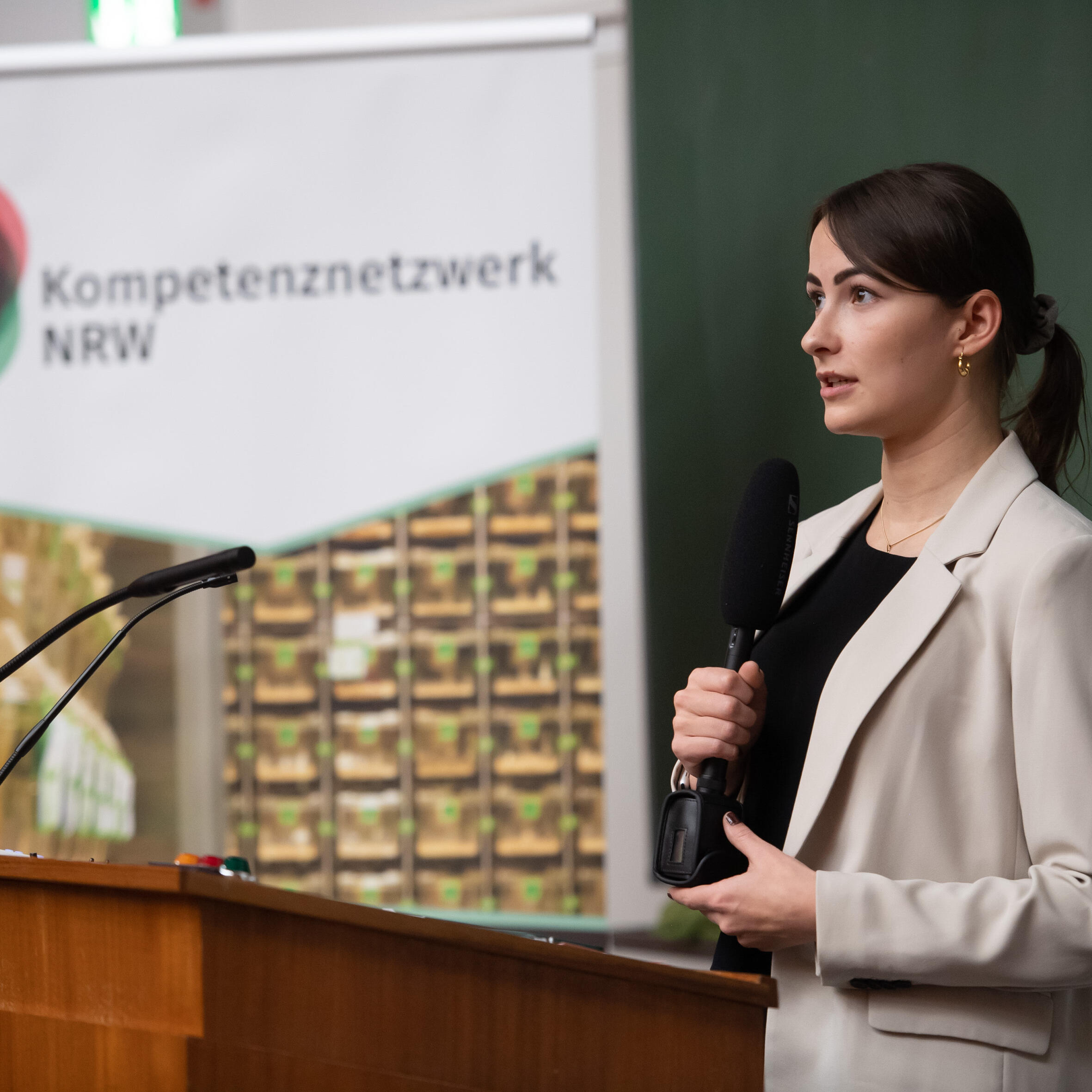 Frau im beigen Blazer und schwarzen Oberteil steht an einem Pult in einem Hörsaal und hält in der rechten Hand ein Mikrofon in der Hand und spricht zum Publikum