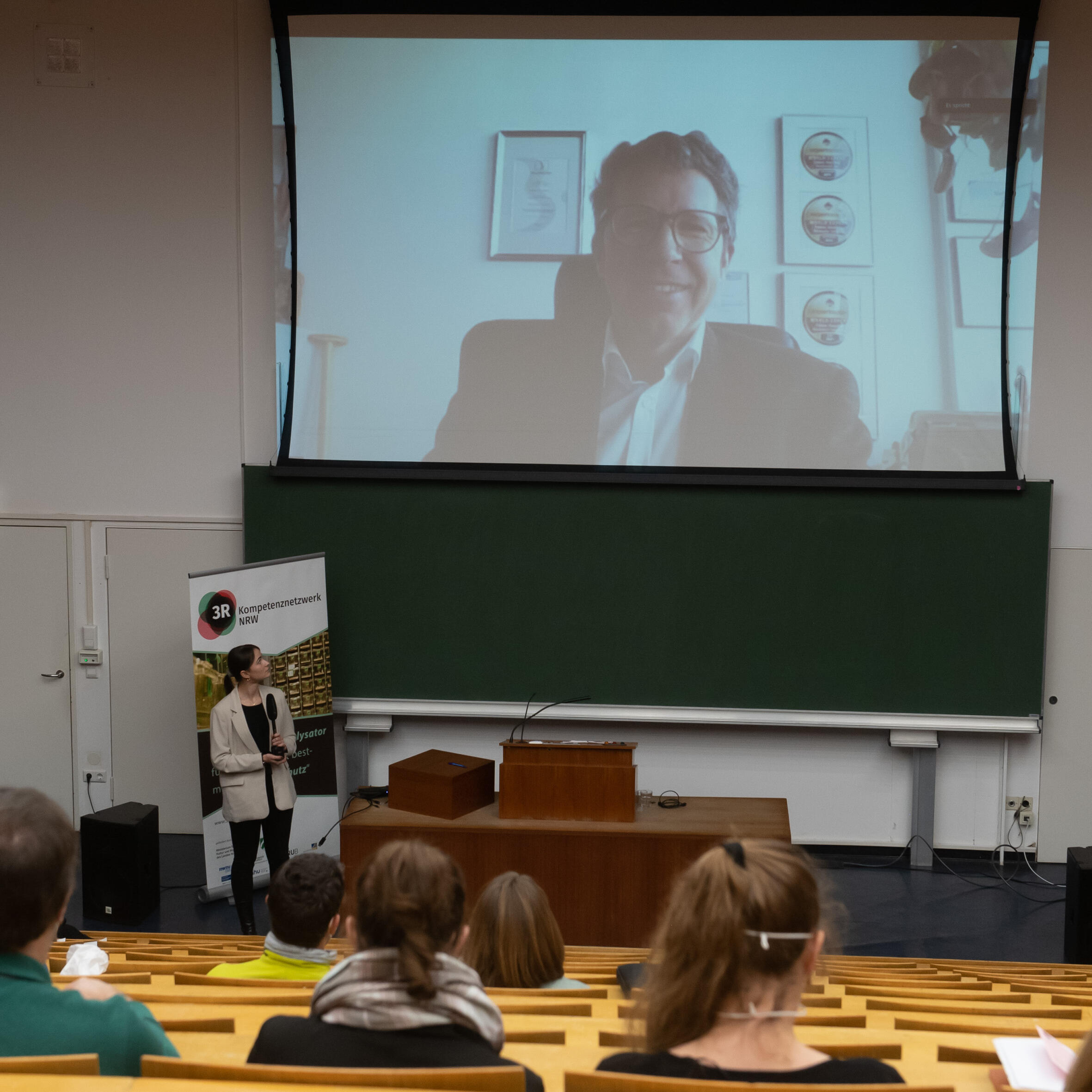 Hörsaal mit Publikum. Alle Blicken auf die Leinwand auf die ein Mann in einem Büro projektziert wird und spricht.