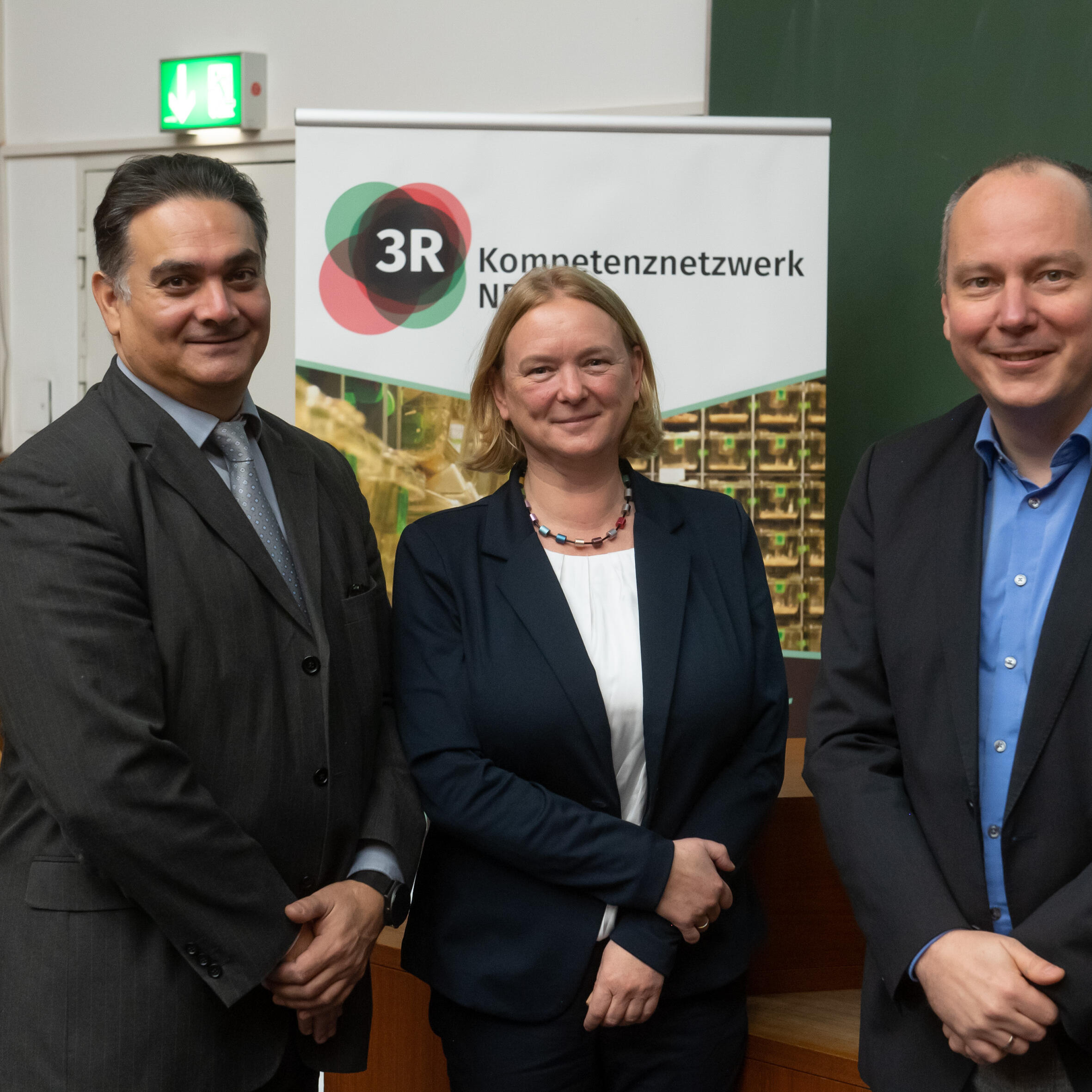 Gruppe von 3 Personen: Linker Mann in einem schwarzgruen Anzug und hellgrauen Hemd, in der Mitte eine Frau mit dunkelblauem Blazer und weißem Oberteil und rechts ein Mann mit schwarzgrauen Anzug und hellblauem Hemd. Alle drei Personen stehen in einem Hörsaal und blicken lächend in die Kamera