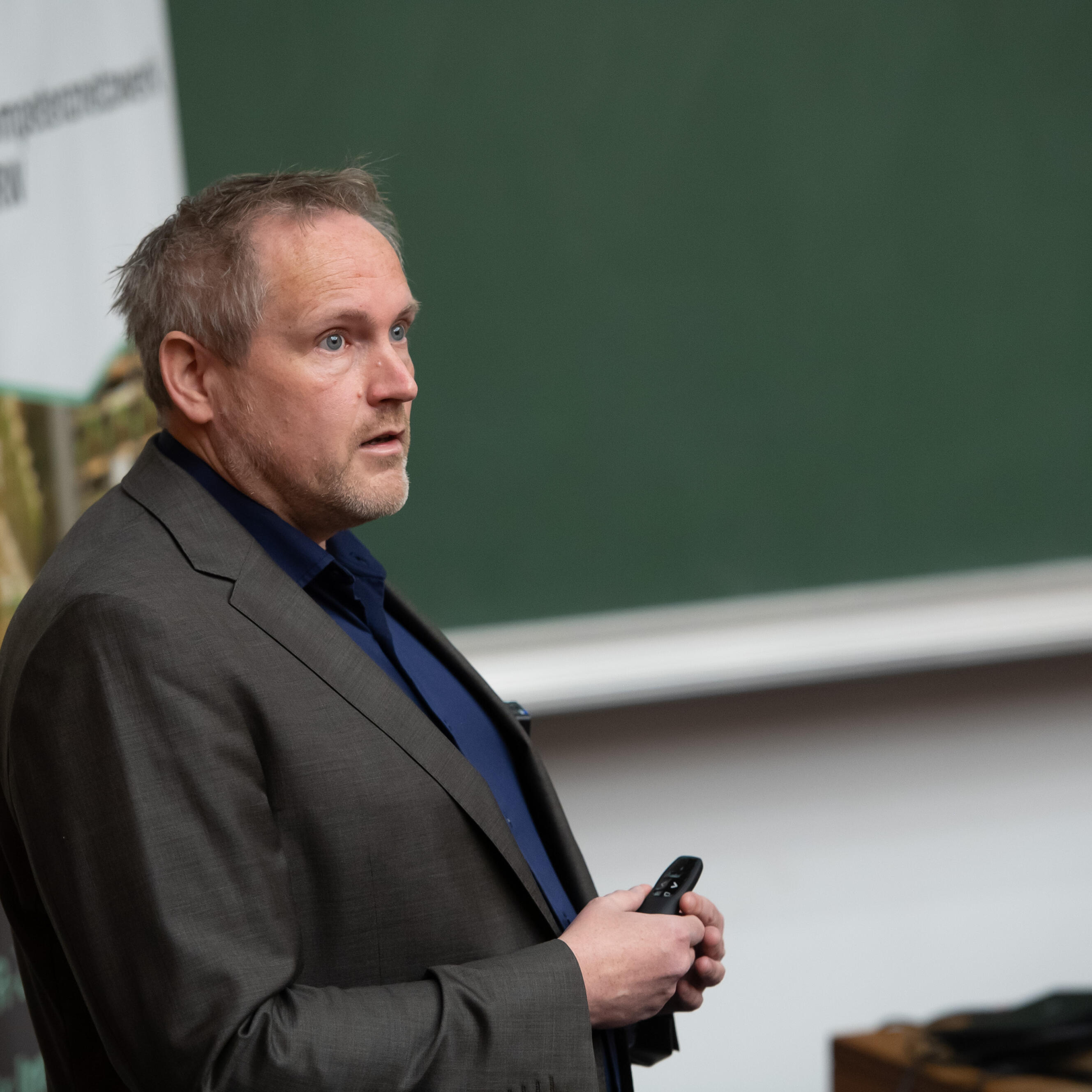 Mann mit grauen Anzug und blauem Hemd steht in einem Hörsaal und blickt das Publikum an. In seiner Hand hält er einen Laserpointer