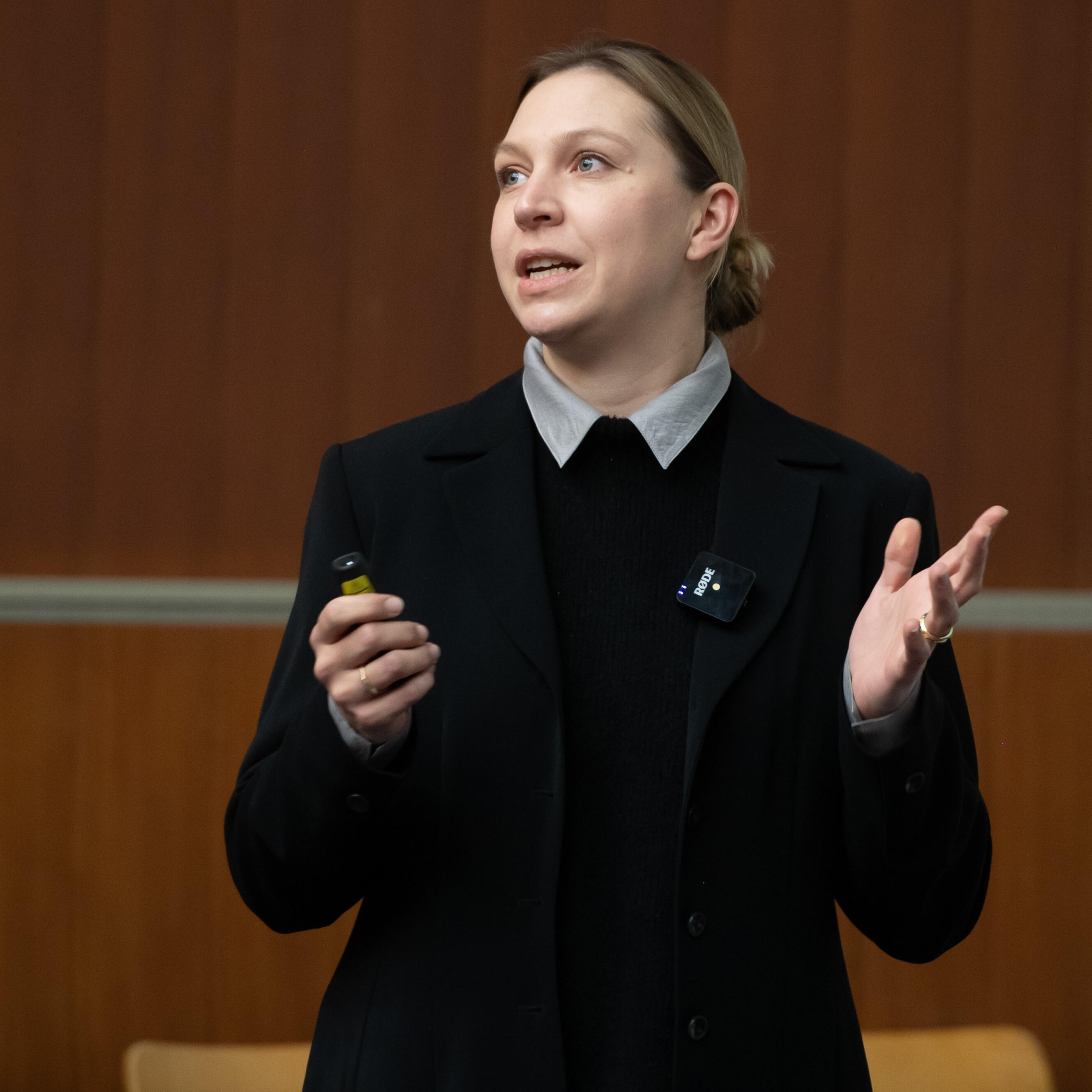 Frau in schwarzen Blazer und schwarzen Oberteil in einem Hörsaal hat den Blick nach links gerichtet und hält in der linken Hand einen Laserpointer und die rechte Hand ist nach vorne zeigend.